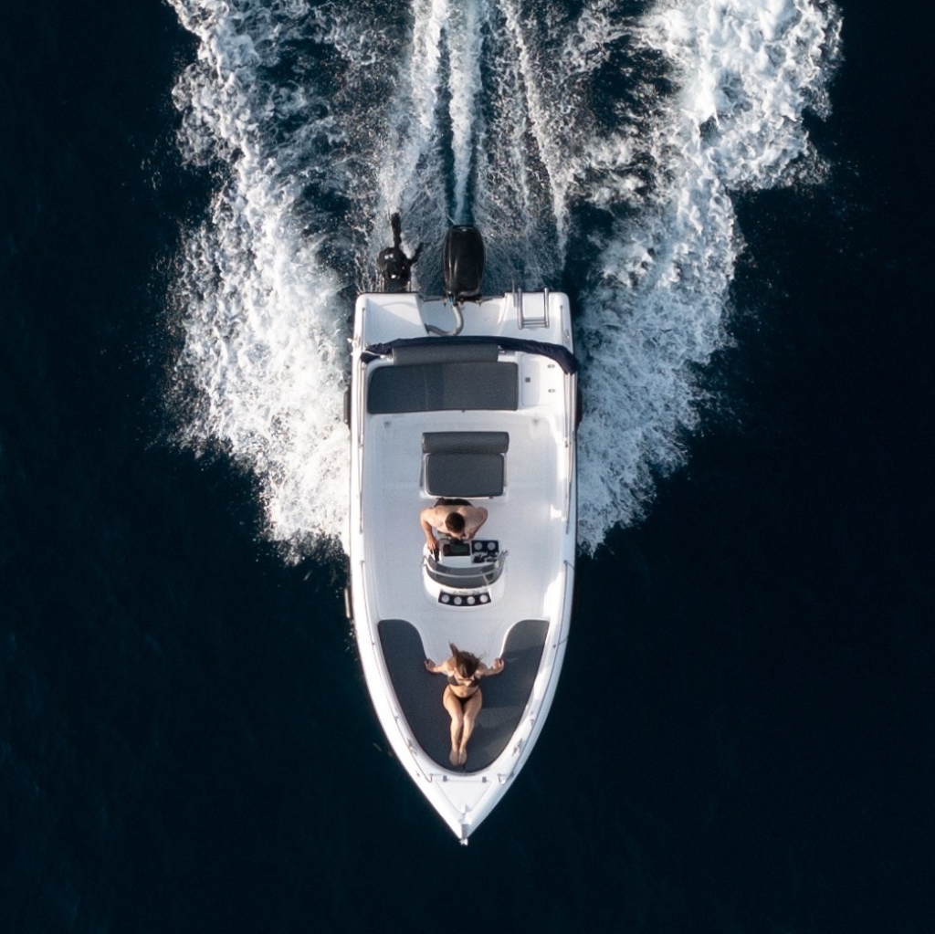 Aerial view of a couple on a self-drive boat rental by Seascape Santorini – woman sunbathing on the bow while partner navigates through the deep blue Aegean Sea. Ideal for romantic private boat tours with no license required.